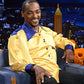Anthony Mackie in a yellow shirt with blue accents sitting on a talk show set with a cityscape background