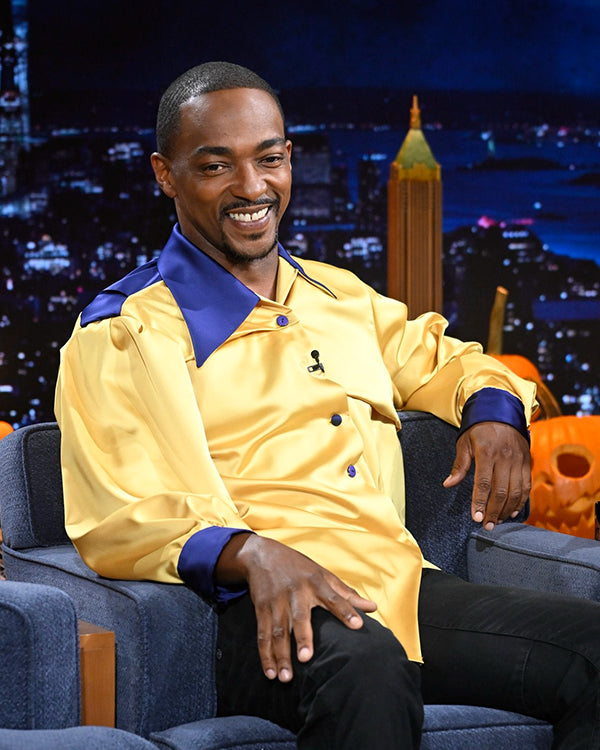 Anthony Mackie in a yellow shirt with blue accents sitting on a talk show set with a cityscape background