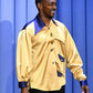 Anthony Mackie wearing a yellow jacket with blue accents against a blue curtain background