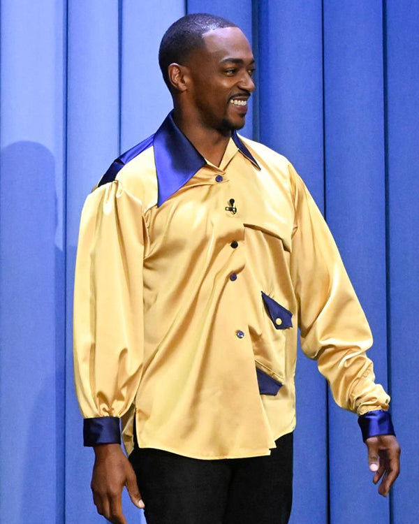 Anthony Mackie wearing a yellow jacket with blue accents against a blue curtain background
